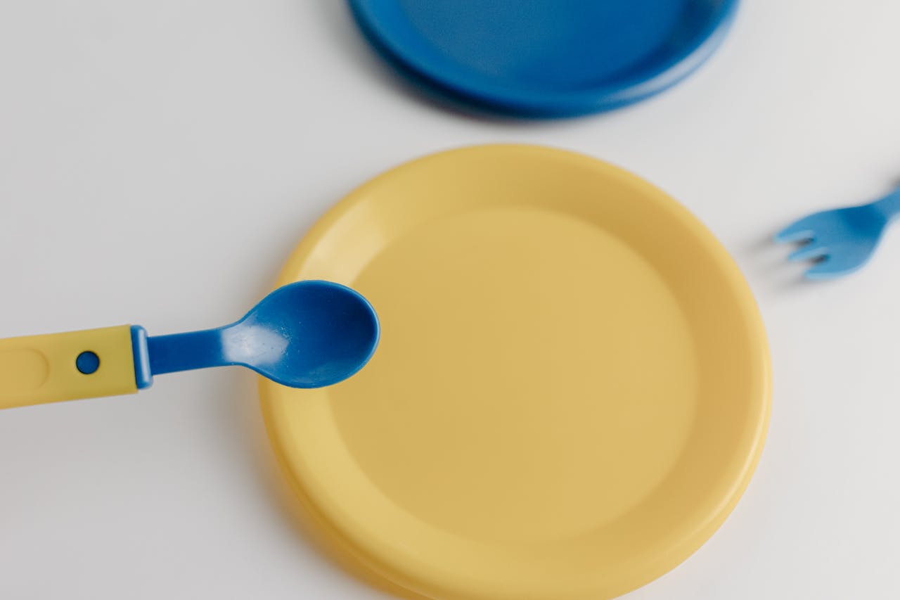 A vibrant plastic toy set featuring a yellow plate, blue spoon, and fork on a white surface.